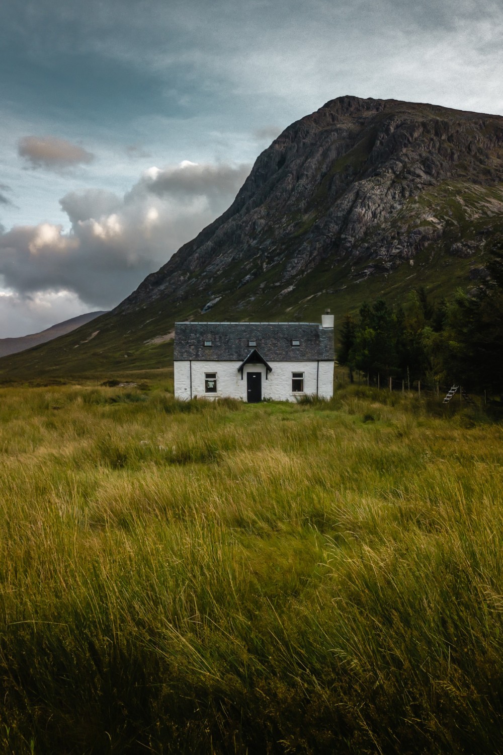 A lone stone house standing in open grassland beneath a mountain ridge